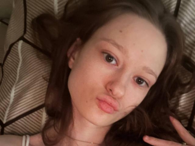 JessieYen has puffy pink lips and brown eyes, lying with wavy brown hair, pale skin, and small moles on her face, wearing a thin necklace, in a relaxed pose.