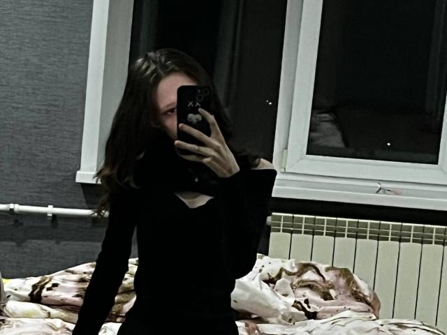 JessieYen with long brown hair, wearing a black long-sleeve top, taking a mirror selfie, covering her mouth with her phone, sitting on a bed with a pink patterned blanket.