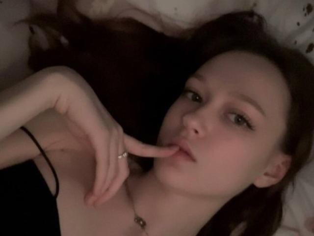 JessieYen's face, fair skin, brown hair, lying down, finger in mouth, black strap, ring on finger, necklace, subtle makeup, slightly parted lips, soft light, relaxed expression.