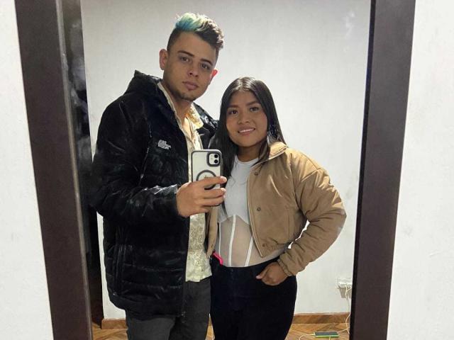 JenniDamian with blue-tipped hair, black puffy jacket, and gray pants, stands next to JenniDamian with long black hair, tan jacket, white shirt, and black pants, smiling.
