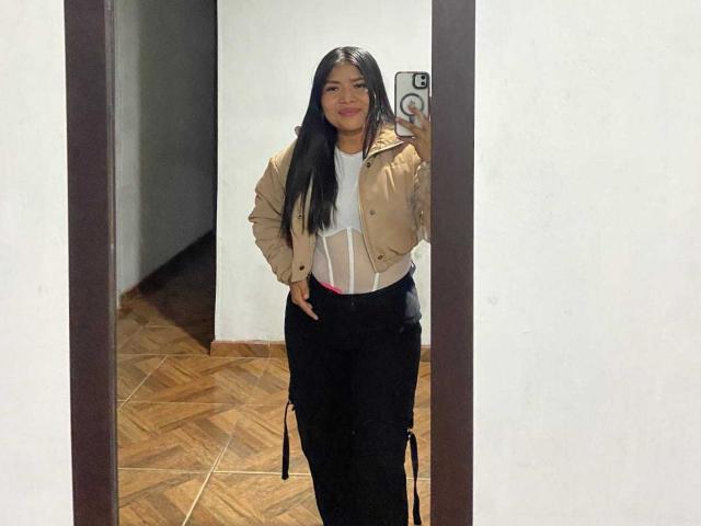 JenniDamian with long black hair, tan jacket, white shirt, and black pants, taking a mirror selfie, hands on hips, smiling, holding a phone with a ring.