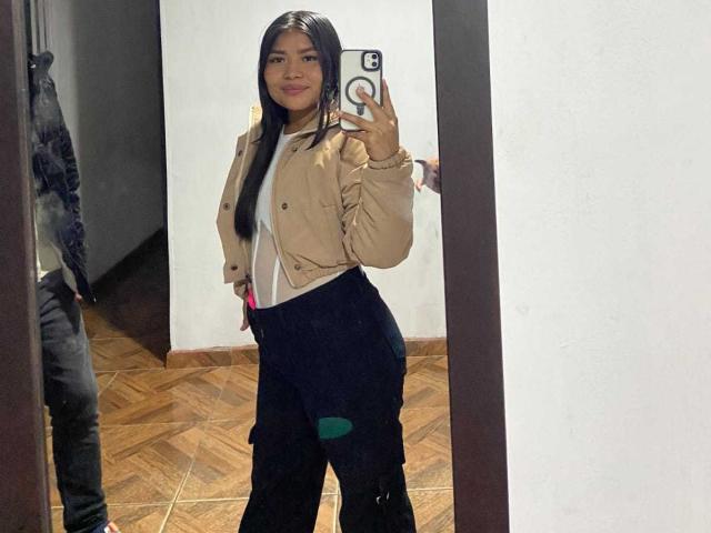 JenniDamian with long black hair, tan jacket, white shirt, black pants, holding phone in mirror selfie, smiling, showing ass, wooden floor, reflection, phone case with ring.