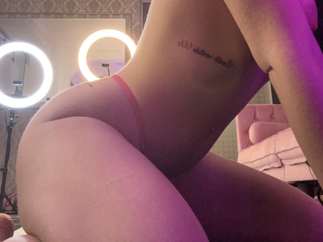 I glutei di AlessiaNoire in un perizoma rosa, un tatuaggio sul fianco, una luce ad anello, una sedia rosa e un'illuminazione viola tenue sullo sfondo. Il suo sedere è il punto focale..