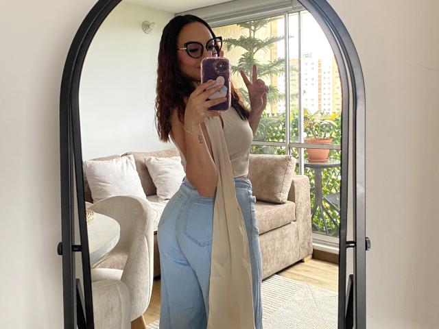 GalaMalkova with wavy red hair, glasses, and a beige top, showing off her ass in high-waisted blue jeans, holding a phone, making a peace sign in a mirror.