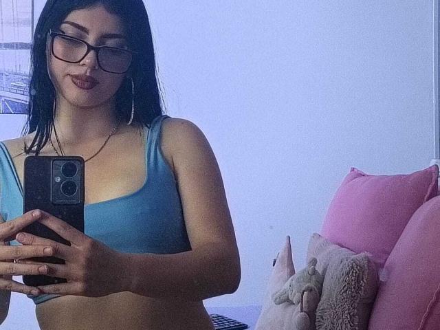 Isabella Rossi mit schwarzen Haaren, Brille und einem blauen Sport-BH, hält ein Telefon, macht ein Selfie und zeigt ihre Brüste. Im Hintergrund sind rosa Kissen und ein Stofftier zu sehen..