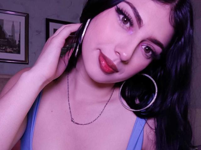 Isabella Rossi, con il trucco degli occhi viola, le labbra rosse lucide, i capelli neri, gli orecchini a cerchio e una maglietta blu, che fa la linguaccia e ha una mano all'orecchio. Il suo seno è piccolo..