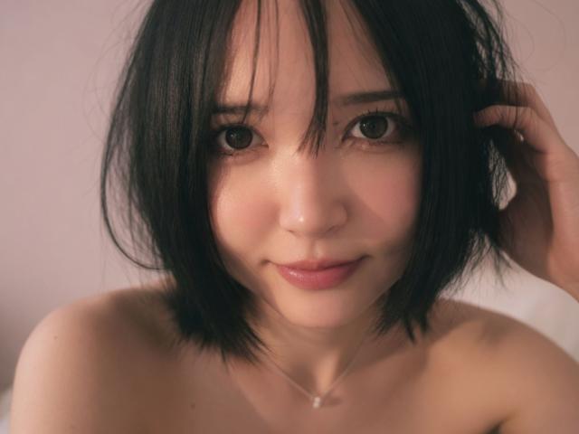 EmaVanhowe has short black hair, brown eyes, and a slight smile. She's topless with a small necklace, her skin is fair, and she's looking directly at the camera.