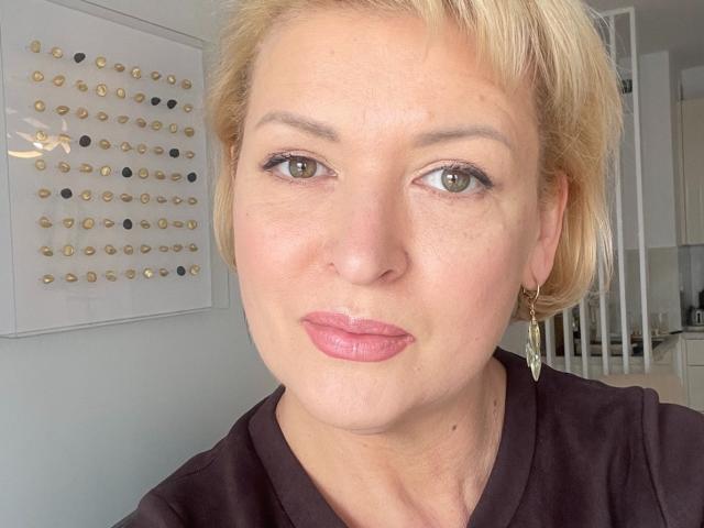 LunaxFantasy has short blonde hair, green eyes, pink lipstick, and wears a black shirt with gold earrings. Her face is slightly flushed. She's looking at the camera.