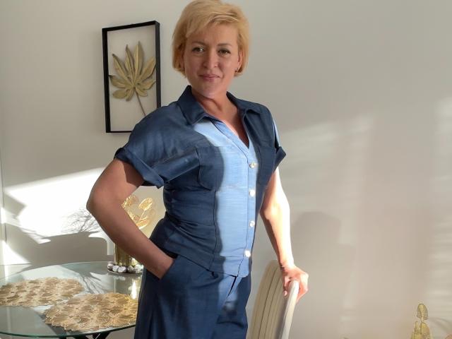 LunaxFantasy heeft kort blond haar, draagt een blauw shirt en broek, staat met haar handen in haar zakken, glimlachend, met zonlicht dat haar borst accentueert en haar borsten laat zien..