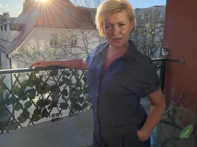 LunaxFantasy mit kurzen, blonden Haaren, trägt ein dunkelblaues Hemd, die Hände in den Taschen, lehnt sich an einen Balkon, die Sonne scheint im Hintergrund, sie wirkt leicht genervt..