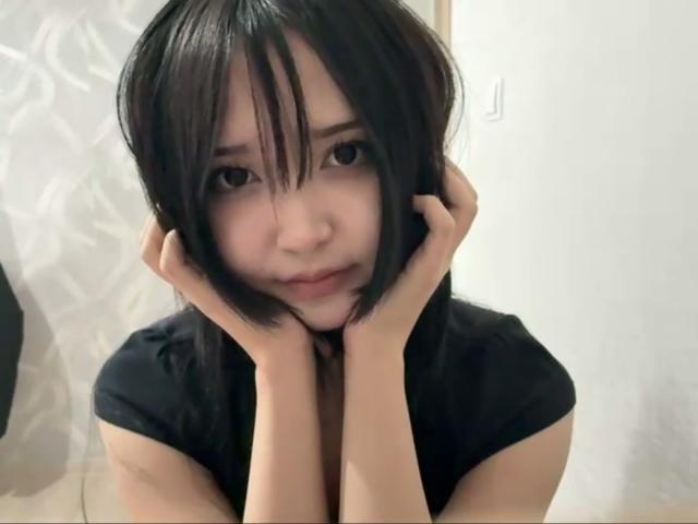 EmaVanhowe has short black hair, hands on her cheeks, looking up with a slight smile. She's wearing a black shirt, and her hands are touching her ears.
