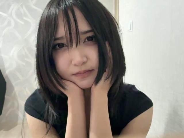 EmaVanhowe with black, shoulder-length hair, hands under chin, black shirt, soft expression, pale skin, looking slightly down, in a casual, indoor setting.