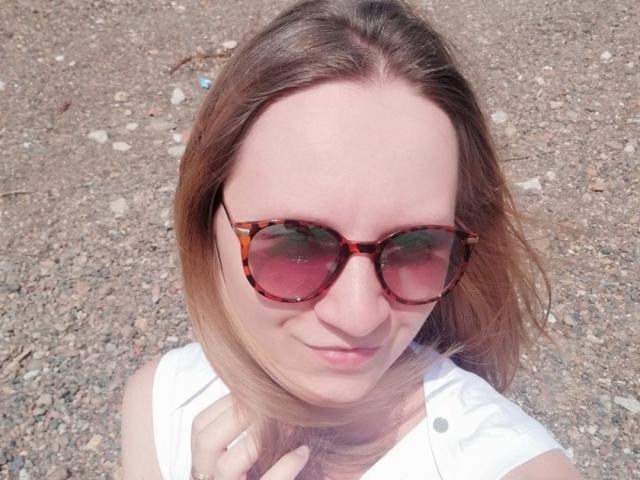 KristalSoetux, con occhiali da sole tartarugati, capelli castano chiaro, una camicia bianca e un leggero sorriso, che tiene la camicia, in piedi su una superficie ghiaiosa..