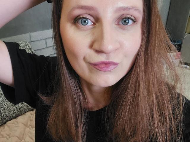 KristalSoetux has blue eyes, pink lipstick, and brown hair, wearing a black shirt, with a slight smile, and a hand in her hair, in a selfie.