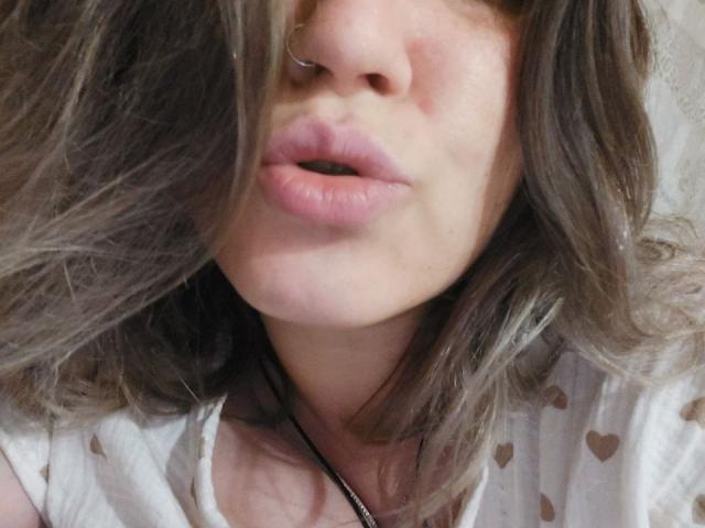 ParadisexWes's face with slightly parted pink lips, brown wavy hair covering one eye, nose ring, and a white shirt with heart patterns. Necklace visible. Soft, natural skin.