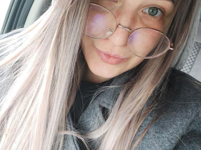 ParadisexWes, con lunghi capelli biondi e lisci, indossa occhiali rotondi rosa e riflettenti, e una giacca grigia con una texture particolare, sorridendo dolcemente. Si vedono le sue labbra rosa e i suoi occhi verdi..