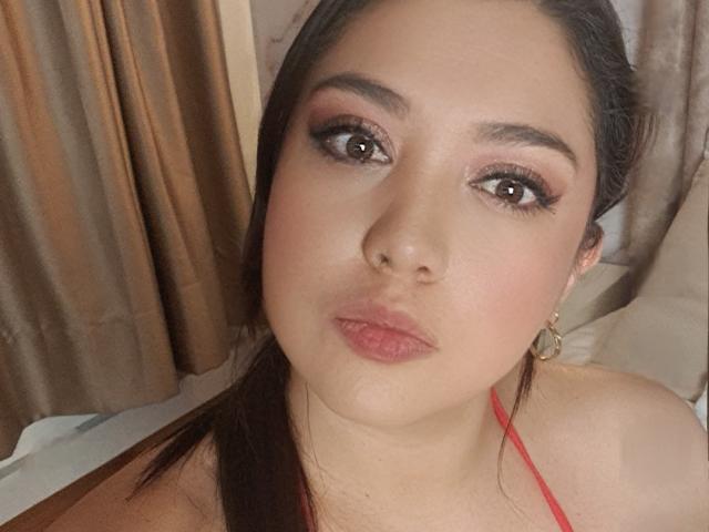 PaulaAnd has brown eyes, full pink lips, and dark brown hair in a ponytail, wearing red earrings and a strap, looking at the camera with a neutral expression.