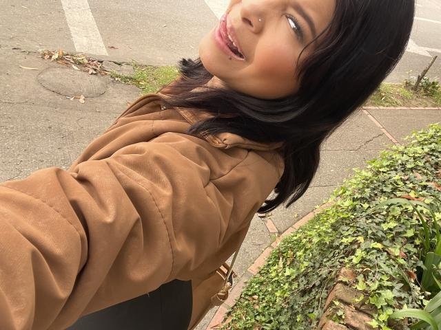 ChloeVixen mit dunklem Haar, einer braunen Jacke und einem Nasenpiercing, lächelt in die Kamera, zeigt ihre perlweißen Zähne und leicht geöffnete, rosafarbene Lippen. Ihre braunen Augen sind halb geschlossen..