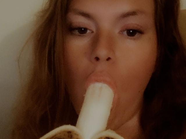 CurvysubAmalia zuigt aan een geschilde banaan, haar lippen omhullen de witte, pluizige punt van de banaan, en haar bruine ogen kijken iets opzij..