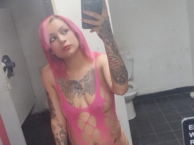 TattedRedd with pink hair, tattoos, and a pink, see-through, fishnet bodysuit, showing her tits and tattoos, taking a selfie in a bathroom. She's holding a phone.
