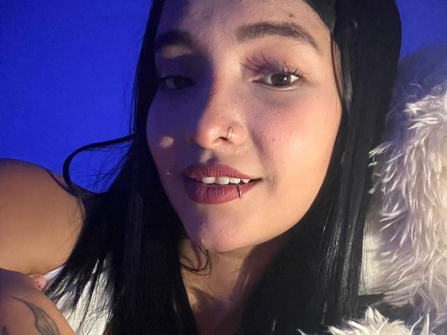 SofiLux has black hair, brown eyes, a nose ring, and a lip piercing with blood on her lower lip. She's smiling with blue lighting and a white furry pillow.