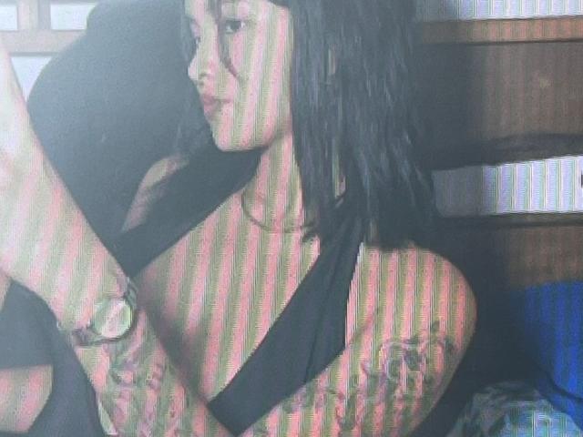 SofiLux with black hair, wearing a black halter top, showing tattooed arms, looking to the side, soft light, blurry, striped shadows on her face and upper body.