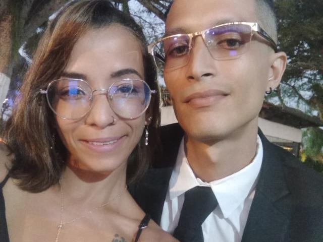 AlejoyMaria with medium-length brown hair, glasses, nose ring, earrings, and a black dress, smiling next to bald AlejoyMaria in a black suit, white shirt, and black tie.