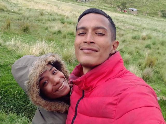AlejoyMaria with medium brown skin, short black hair, in a red puffy jacket, smiling. AlejoyMaria's dark-haired, brown-skinned friend with a furry hood, smiling, leans on AlejoyMaria's shoulder.