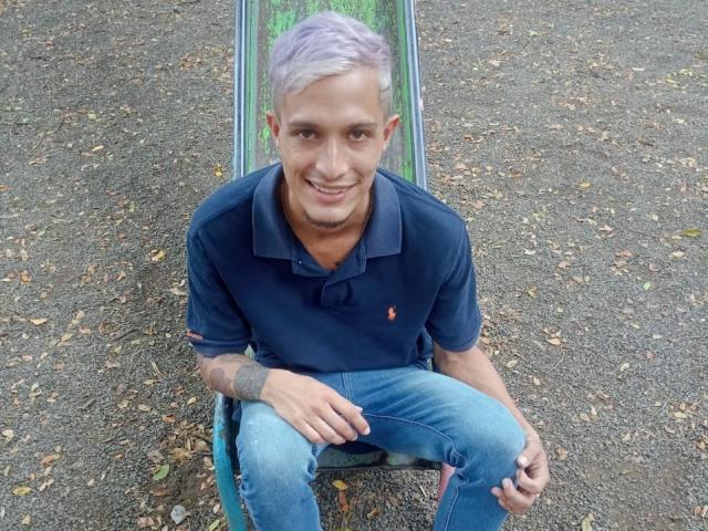 AlejoyMaria with short purple hair, wearing a navy polo and blue jeans, sitting on a skateboard, smiling, tattoo on right forearm, gravel ground, hands resting on knees.