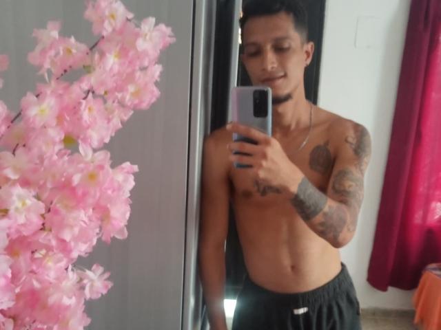 Shirtless AlejoyMaria with tattoos, holding phone, pink cherry blossom branch in mirror, black choker, black pants, short black hair, slight smile, right arm tattoo, red curtain background.