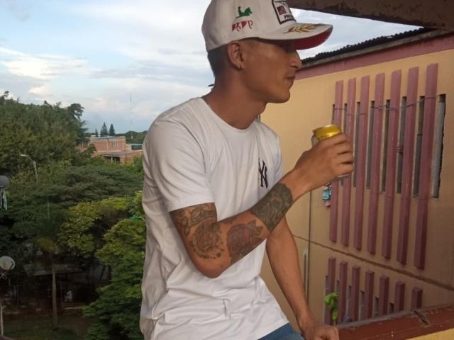 AlejoyMaria, wearing a white NY cap and shirt, sips from a yellow cup, showing tattoos on his right arm, standing on a red-tiled balcony, looking to the side.