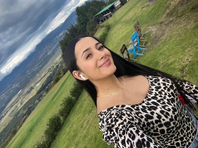 IsabellaAustin with long black hair, smiling, wearing a leopard print top, white earbuds, and a gold necklace, standing in a grassy field with mountains and chairs in background.