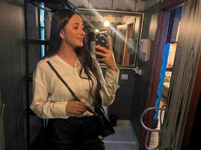IsabellaAustin with long brown hair, white shirt, black bag, holding phone, smiling, showing her tits in mirror selfie, reflection showing light bulb.