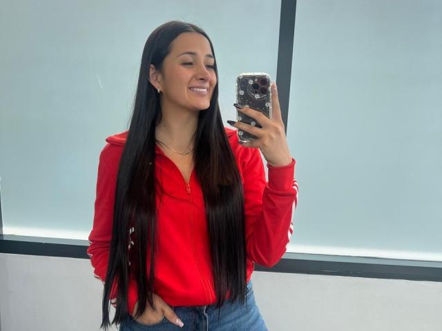 IsabellaAustin with long black hair, red hoodie, and blue jeans, smiling while taking a selfie with a phone covered in a black and white pattern. Hands in pockets.