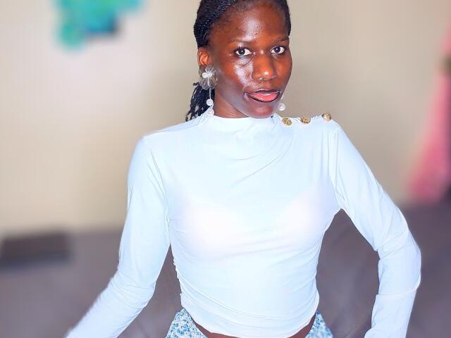 KharlaQueen with dark skin, wearing a white long-sleeve top showing her small tits, standing with hands on hips, and a blue patterned skirt, in a blurred background.