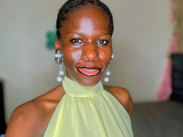 KharlaQueen with dark skin, shiny face, and black braided hair, wearing a sheer yellow halter top, and shiny pink lipstick, smiling with her ass covered by the top.