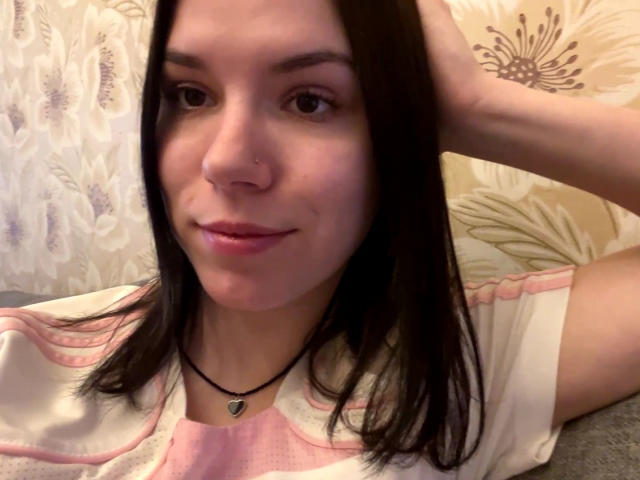MagdalenaMurchie with straight dark brown hair, small nose ring, pink lipstick, white shirt with pink accents, black necklace, and a subtle smile, resting hand on head.