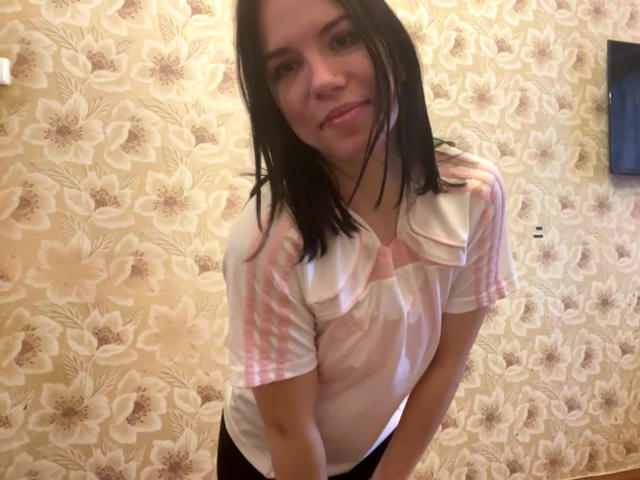 MagdalenaMurchie with wet black hair, wearing a pink and white striped shirt, leaning forward, smiling, showing small tits, and a slim ass, against floral wallpaper.