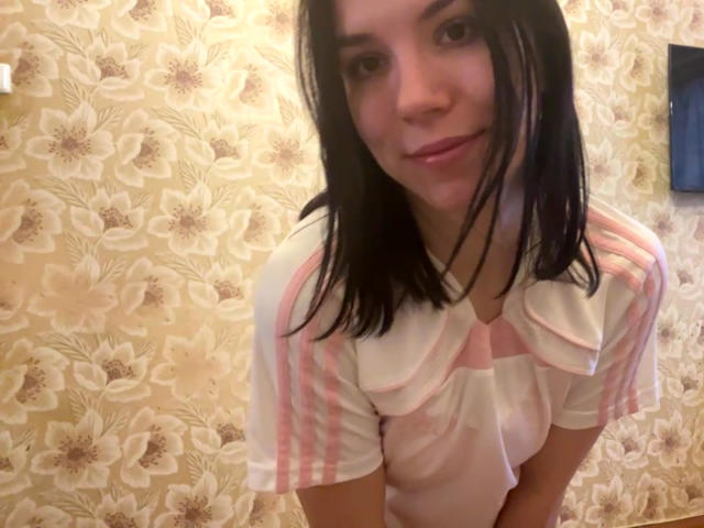 MagdalenaMurchie with medium tits, wearing a pink and white striped shirt with a ruffled collar, black hair, slight smile, standing in a room with floral wallpaper.