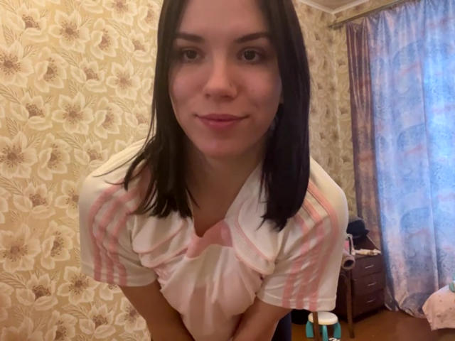 MagdalenaMurchie with black hair, pink and white striped shirt, leaning forward, smiling, showing small tits, in a room with floral wallpaper.
