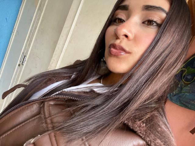 MilaDreams with glossy brown lips, straight brown hair, brown leather jacket, white shirt, and nose ring, looking at camera with subtle smile, showing her upper body.