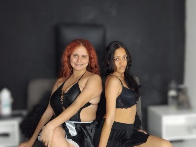 Kiara e Gloria, con capelli rossi ricci e grandi seni, indossano un reggiseno di pizzo nero e una gonna di seta, sedute una di fronte all'altra su una sedia. Gloria, con i capelli neri e piccoli seni, indossa un reggiseno di pizzo nero..