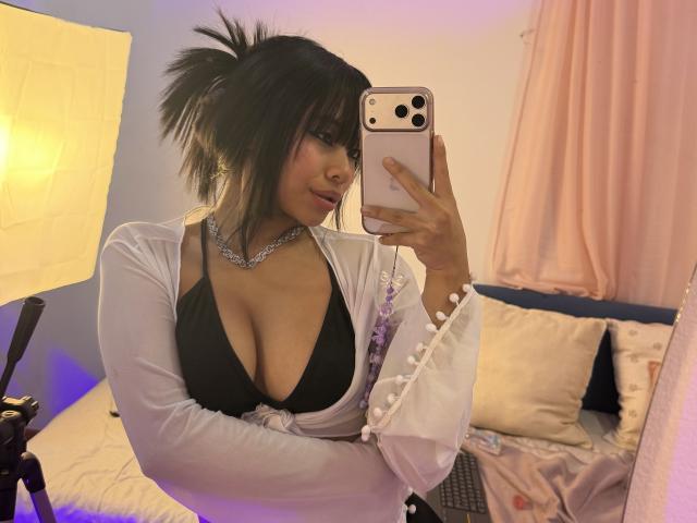 VannesaSaenz with dark hair in a messy ponytail, wearing a white cardigan, black deep cleavage, and silver necklace, taking a mirror selfie with her phone. Boobs visible.