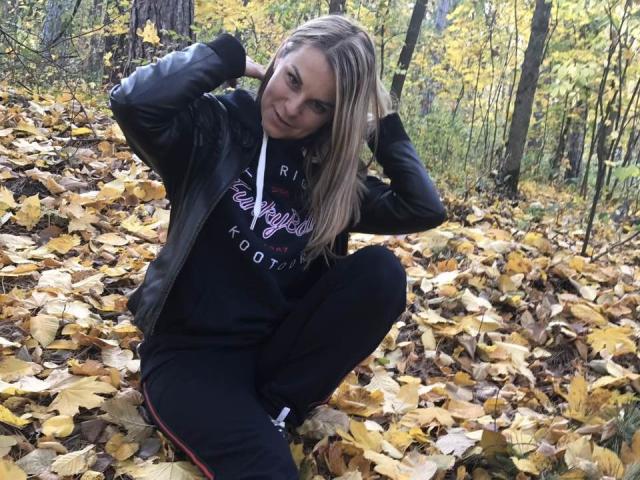 BelleLisaG avec des cheveux blonds, portant une veste en cuir noir et un pantalon noir, assise sur des feuilles d'automne jaunes, les mains dans les cheveux, souriante, avec un arrière-plan de forêt..