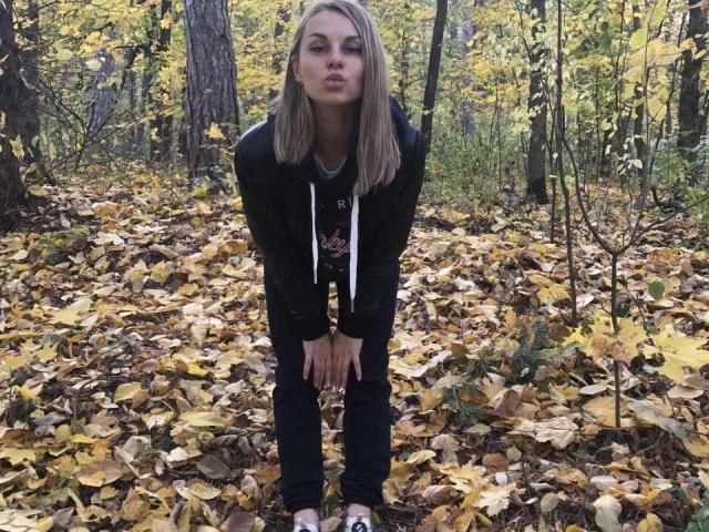 BelleLisaG with blonde hair, black jacket, and dark jeans, pouting in a forest covered in yellow autumn leaves, hands on knees, wearing a gray shirt.