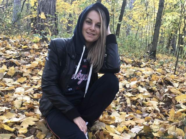 BelleLisaG with blonde hair, black hoodie, and leather jacket, squatting on yellow autumn leaves, smiling, hand in hair, black pants, forest background, natural light, casual, cute.