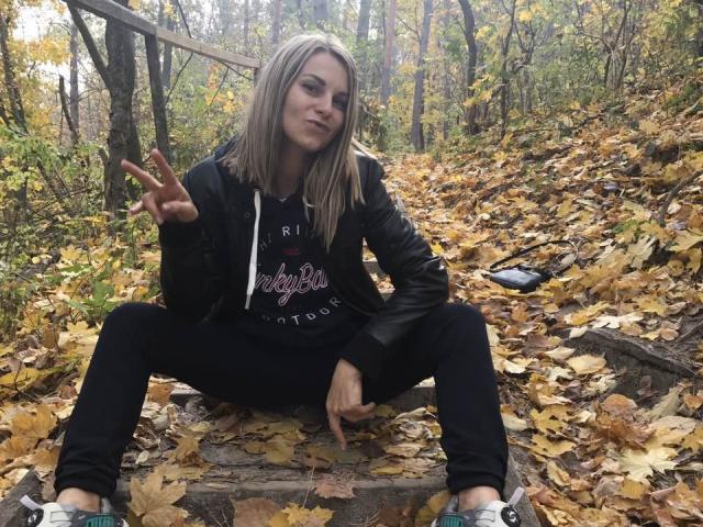 BelleLisaG, blonde, leather jacket, black hoodie, sitting on steps, spreading legs, making peace sign, autumn leaves, black pants, sneakers, forest background, relaxed, confident, cute.