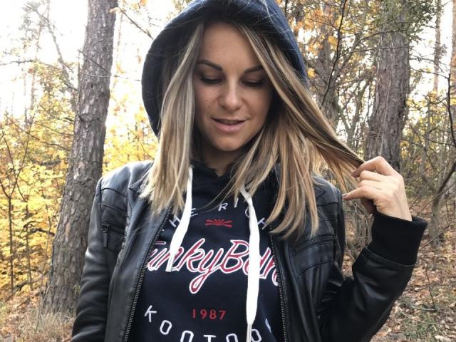 BelleLisaG with blonde hair, wearing a black leather jacket and blue hood, gently pulling her hair, smiling, in a forest with yellow autumn leaves.