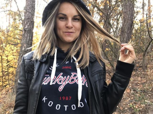BelleLisaG, avec ses cheveux blonds, sa veste en cuir noir et son t-shirt noir à capuche, tenant ses cheveux avec un léger sourire, sur fond de forêt d'automne. Ses seins sont partiellement couverts par le t-shirt..