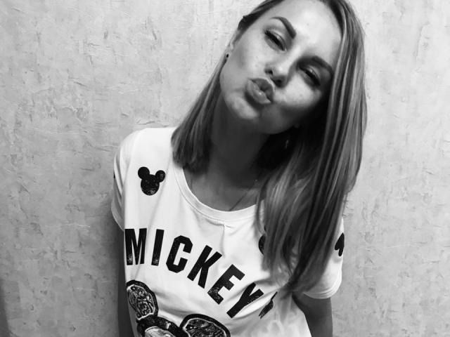 BelleLisaG with pouty lips, slightly tilted head, and straight shoulder-length hair, wearing a white "MICKEY" shirt, black bow, against textured wall, in black-and-white photo.